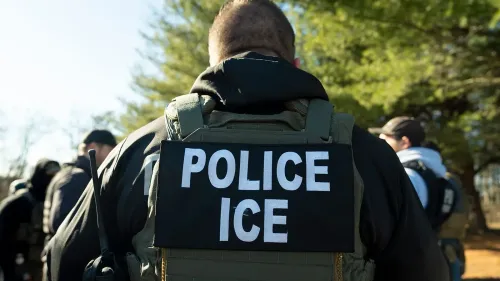 ضابط إنفاذ قانون يرتدي سترة مكتوب عليها "شرطة ICE"، في سياق تنفيذ قوانين الهجرة في الولايات المتحدة، مع ظهور أفراد آخرين في الخلفية.