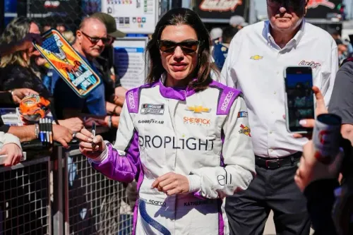 كاثرين ليج، سائقة سباقات بريطانية، ترتدي بدلة سباق وتوقع تذكارات للمعجبين خلال حدث سباق ناسكار، محاطة بجمهور.