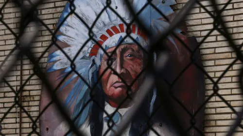 صورة لجدارية تُظهر وجه هندي أمريكي يرتدي غطاء رأس مزخرف، محاطة بسياج معدني، تعكس الجدل حول تمائم الفرق الرياضية.