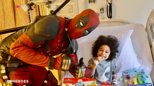 ديدبول يرتدي زيه الشهير ويحتفل مع طفل مبتسم في المستشفى، محاطاً بالألعاب والهدايا، مما يعكس لحظة سعادة وبهجة.