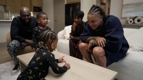 عائلة مختلطة الثقافات تتفاعل في غرفة معيشة دافئة، حيث يلعب طفل على الطاولة بينما يراقبه البالغون بابتسامات محبة.