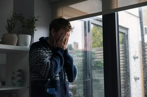 رجل يرتدي سترة داكنة، يغطي وجهه بيديه، ويبدو متوترًا أمام نافذة مبللة بالمطر، مما يعكس مشاعر القلق والإجهاد.