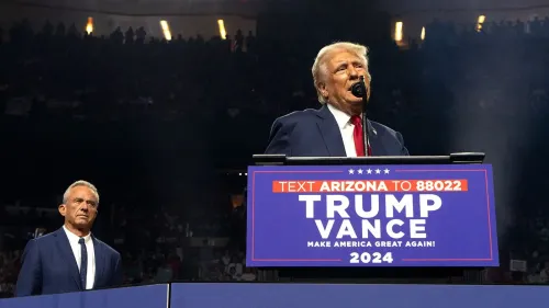 دونالد ترامب يتحدث في تجمع انتخابي بينما يقف روبرت كينيدي الابن خلفه، مع لافتة تدعو لدعمهما في الانتخابات.