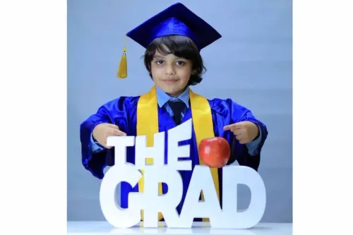 طفل يرتدي قبعة التخرج وزيّاً أكاديمياً، يشير بفخر إلى كلمة "THE GRAD" مع تفاحة حمراء، معبراً عن فرحة التخرج والإنجاز.