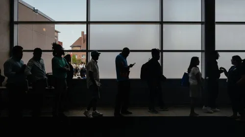 طابور من الأشخاص ينتظرون في مكان داخلي، مع إضاءة خلفية تبرز ظلالهم، مما يعكس تأثيرات سوق العمل والبطالة في الولايات المتحدة.