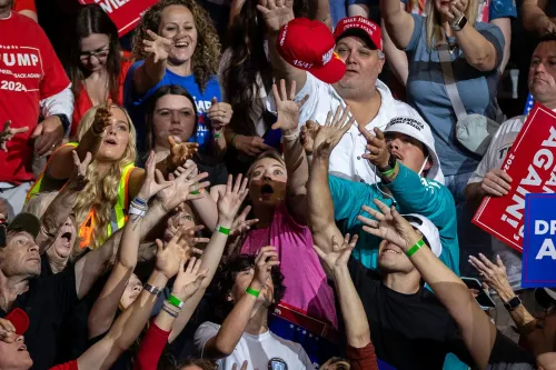 حشد من المؤيدين في تجمع سياسي، يمدّون أيديهم نحو قبعة تحمل شعار ترامب، مع لافتات تدعم حملته الانتخابية لعام 2024.