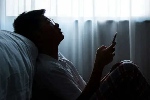 شخص يجلس على السرير، يحمل هاتفه الذكي، مع إضاءة خافتة من النافذة، يعكس شعور القلق والتفاعل مع وسائل التواصل الاجتماعي.