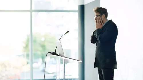 رجل يرتدي بدلة يقف أمام منصة، يبدو عليه التوتر والقلق، مستعدًا للتحدث أمام الجمهور في موقف ضغط.