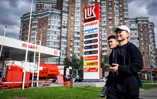 شبان يمشيان بالقرب من محطة وقود "لوك أويل" مع لافتة تعرض أسعار الوقود، تعكس تأثيرات الحرب على الاقتصاد الروسي.