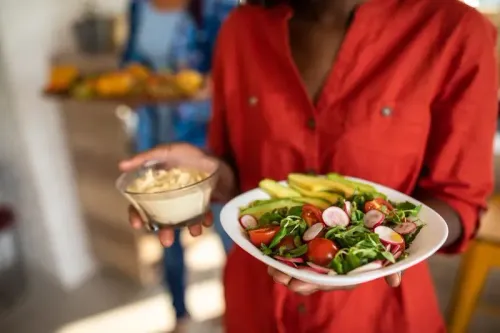 امرأة تحمل طبقًا من السلطة الطازجة مع الخضروات، بينما تمسك بيدها الأخرى وعاءً صغيرًا من الحمص، مما يعكس أهمية التغذية الصحية في الوقاية من سرطان القولون والمستقيم.