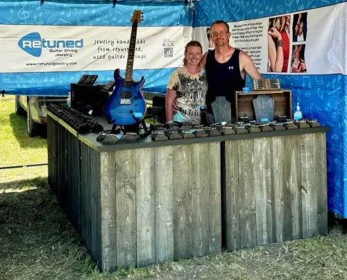 زوجان يقفان أمام طاولة عرض مجوهرات مصنوعة من أوتار الغيتار المعاد تدويرها، مع لافتة لمشروعهما "ريتونيد للمجوهرات".