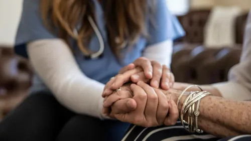 تظهر الصورة يدي ممرضة وهي تمسك بيد امرأة مسنّة، مما يرمز إلى الرعاية والدعم في مراكز رعاية المسنين.
