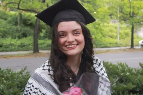 فتاة ترتدي قبعة تخرج وتبتسم، تحمل باقة من الزهور، وتظهر خلفها أشجار خضراء، تعبيرًا عن الفرح والنجاح.