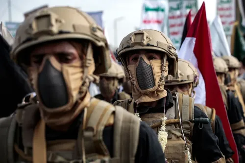 جنود يرتدون ملابس عسكرية وأقنعة، يشاركون في مظاهرة، مع أعلام خلفهم، في سياق الأزمة الإنسانية في اليمن وتأثير الحوثيين.