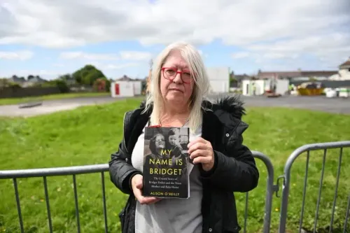 امرأة تحمل كتاباً بعنوان "اسمي بريدجيت" في موقع التنقيب عن مقابر جماعية لأطفال في توام، أيرلندا، حيث تم اكتشاف رفات حوالي 800 رضيع.
