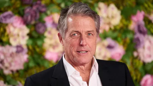 يظهر هيو غرانت مبتسمًا في حدث رسمي، مع خلفية زهور ملونة، مما يبرز أجواء الحفل.