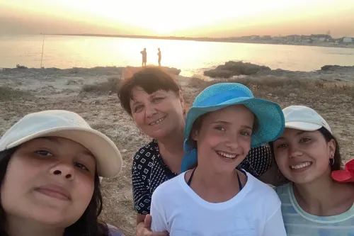 صورة لثلاث فتيات وامرأة مسنّة على شاطئ، تبتسم الفتيات في مقدمة الصورة بينما يظهر غروب الشمس في الخلفية، مما يعكس لحظة من السعادة وسط الأوقات الصعبة.
