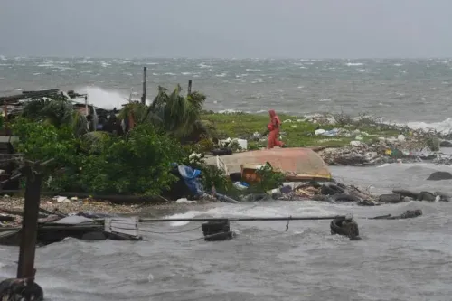تظهر الصورة شخصًا يرتدي ملابس واقية في منطقة تضررت من إعصار ميليسا، مع وجود حطام ومياه مرتفعة على الشاطئ.