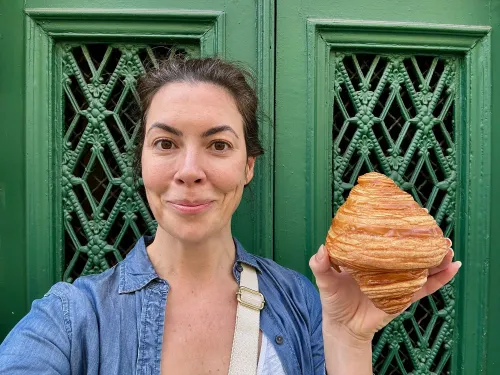 امرأة تحمل كرواسون على شكل حلزوني أمام باب أخضر مزخرف، تعبر عن سعادتها بحياتها الجديدة في أوزيس، فرنسا.