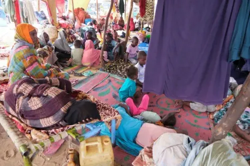 مخيم للنازحين في السودان، حيث يجلس الناس على الأرض في ظل خيام بسيطة، مع أطفال يلعبون، مما يعكس الأوضاع الإنسانية الصعبة.
