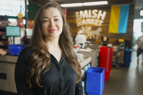 امرأة ترتدي سترة سوداء وقميص ICE، تخفي عينيها بنظارات شمسية وقناع، تتحدث مع موظف في فندق، وسط أجواء توتر.