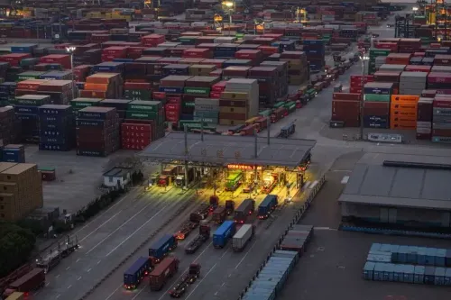 مستودع حاويات كبير يضم شاحنات متوقفة، مع تكدس الحاويات الملونة، يعكس تأثير التجارة العالمية على الاقتصاد.