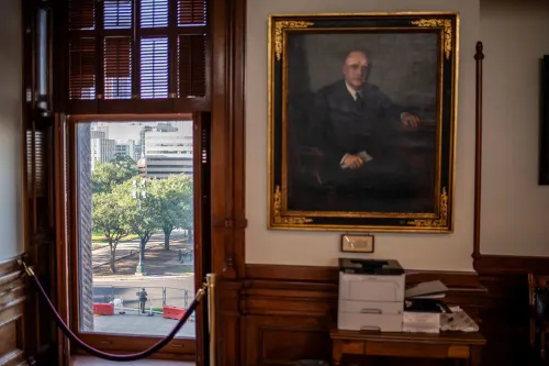 منظر من نافذة مكتب يظهر حديقة خارجية مع أشجار، بالإضافة إلى لوحة فنية لرجل يرتدي بدلة، تعكس أجواء السياسة في تكساس.