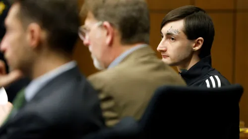 رجل متهم بإطلاق نار جماعي يجلس في قاعة المحكمة، محاطًا بمحاميه، حيث تبدأ محاكمته في قضية مأساوية تتعلق بإطلاق النار في موكب عيد الاستقلال.