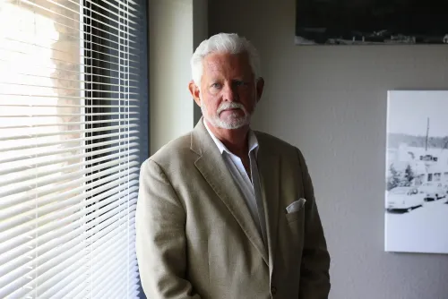 رجل مسن ذو لحية وشعر أبيض يقف بجانب نافذة مع ستائر، يرتدي بدلة رمادية، في خلفية تحتوي على صورة بالأبيض والأسود.