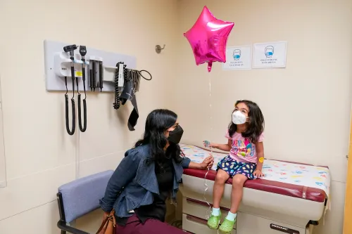 طفلة ترتدي قناع وجه وتجلس على سرير طبي في عيادة، تحمل بالونًا ورديًا، بينما تجلس والدتها بجانبها.