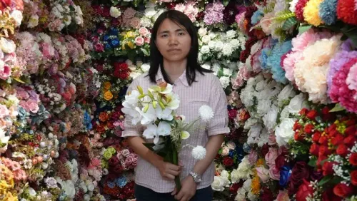 امرأة تحمل زهورًا صناعية في سوق ييوو الصيني، المحوري في تجارة زينة عيد الميلاد، وسط خلفية ملونة من الزهور.