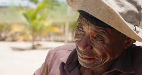 وجه رجل مسن يرتدي قبعة، يظهر عليه الحزن والتعب، في خلفية خضراء. تعكس ملامحه تأثير الصراع والفقر في موزمبيق.