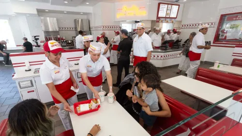 مطعم In-N-Out يقدم خدمة للزبائن مع موظفين يرتدون زي العمل، ويظهر في الخلفية طاولات حمراء ومطبخ مفتوح.