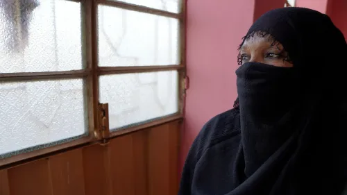 امرأة ترتدي الحجاب، تنظر من نافذة في غرفة ذات جدران ملونة، تعكس معاناة الناجيات من العنف الجنسي في السودان.