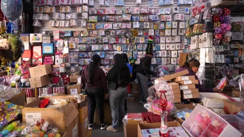 متسوقون يستعرضون مجموعة متنوعة من البضائع في متجر مزدحم، مع حائط مغطى بالبطاقات والهدايا، مما يعكس تأثير الرسوم الجمركية على الأسعار.