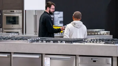 شخصان يتحدثان في متجر للأجهزة المنزلية، مع وجود معدات مطبخ حديثة في الخلفية، مما يعكس اتجاهات السوق الحالية.