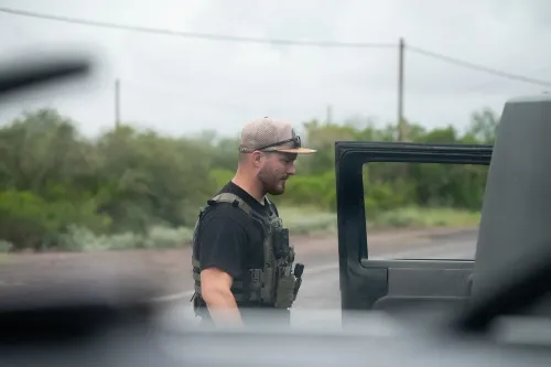 رجل يرتدي سترة واقية وقبعة، يقف بجانب سيارة في منطقة صحراوية، في سياق تحقيق حول تهريب المهاجرين.