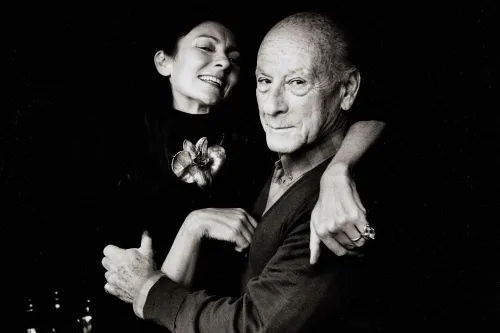 صورة بالأبيض والأسود لامرأة مبتسمة تحتضن رجلًا مسنًا، مع خلفية داكنة، تعكس العلاقة الحميمية بينهما في عالم الفن.