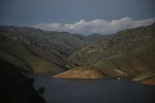 منظر طبيعي يُظهر بحيرة محاطة بالتلال الخضراء تحت سماء زرقاء مع سحب خفيفة، تعكس تأثيرات إدارة المياه في كاليفورنيا.