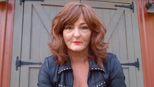 امرأة ذات شعر بني مموج وحواجب مرتفعة، ترتدي سترة جلدية، تظهر في المقدمة مع باب خشبي خلفها.