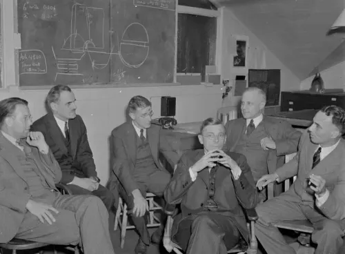 اجتماع لمجموعة من العلماء في غرفة دراسية، يناقشون أبحاثًا علمية، مع وجود لوح تعليمي في الخلفية.