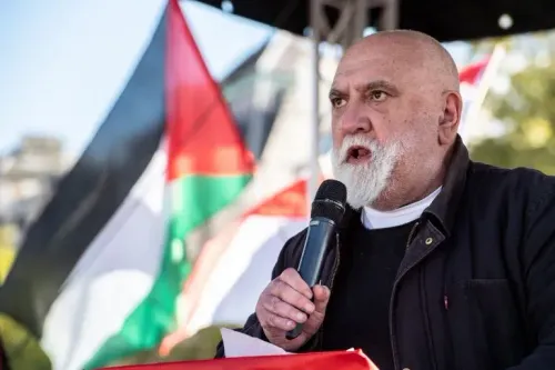 رجل مسن يتحدث في تجمع احتجاجي، محاطًا بأعلام فلسطينية، معبرًا عن دعمه لحقوق الفلسطينيين.