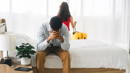 رجل يجلس حزينًا على حافة السرير بينما تتركه امرأة تحمل حقيبة سفر، مما يعكس مشاعر الانفصال أو الطلاق.