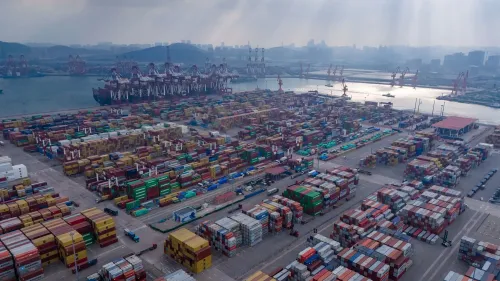 صورة لميناء شحن مزدحم يحتوي على حاويات ملونة، مع سفن وشاحنات، تعكس تأثير الرسوم الجمركية على التجارة الأمريكية.