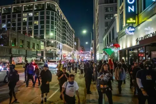 احتجاجات في لوس أنجلوس ليلاً، حيث يسير المتظاهرون في الشارع، مع وجود علم مكسيكي يرفرف في الخلفية، وسط أضواء المدينة.