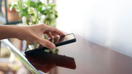 يد تظهر يدًا تلامس هاتفًا ذكيًا موضوعًا على طاولة، مع نباتات خضراء في الخلفية، مما يعكس الاعتماد على وسائل التواصل الاجتماعي.