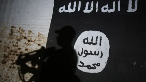 ظلّ شخص مسلح يحمل سلاحًا، مع ظهور ظله على جدار يحمل شعارات لتنظيم الدولة الإسلامية، مما يعكس التوتر الأمني في العراق.
