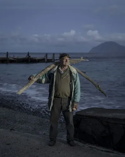 رجل يحمل عصا كبيرة على كتفه، واقفاً على شاطئ جزيرة أليكودي الهادئة، مع مناظر طبيعية بركانية جيئة وذهاباً في الخلفية.