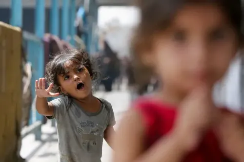 طفلة صغيرة ترتدي قميصًا رماديًا، تعبر عن مشاعرها في مخيم للاجئين، بينما تظهر طفلة أخرى في الخلفية. تعكس الصورة معاناة الأطفال في غزة.