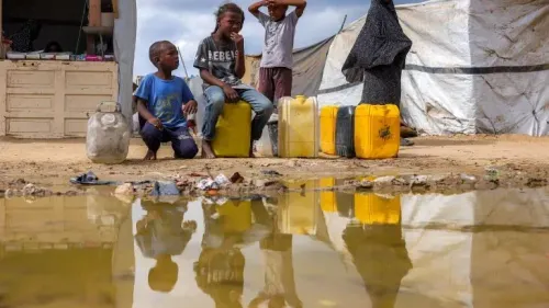 أطفال يجلسون بجوار حاويات مياه في مخيم للنازحين، يعكسون معاناة الحياة اليومية في غزة وسط ظروف إنسانية صعبة.